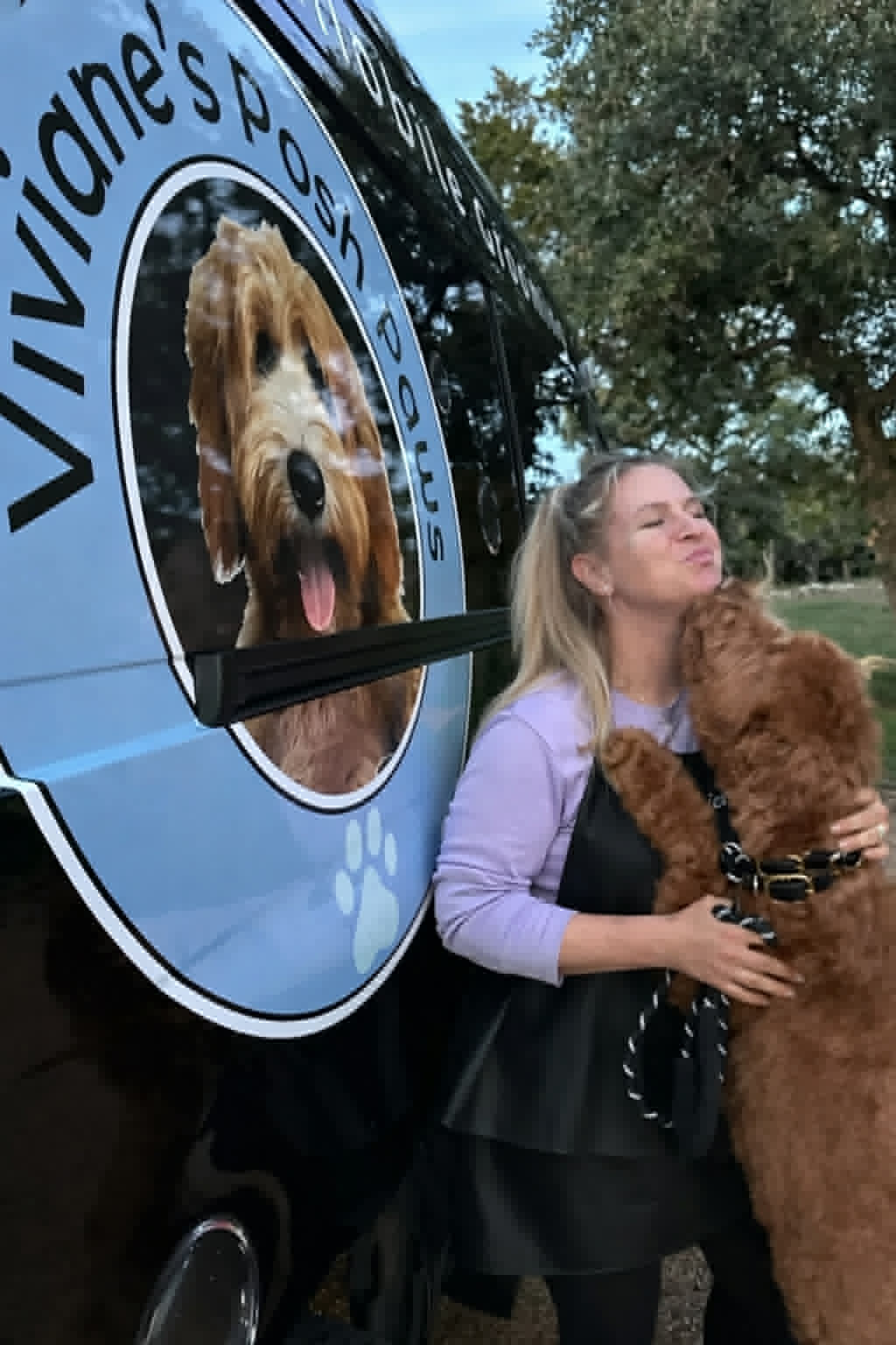 A groomer hugging a red doodle beside the Viviane's Posh Paws mobile grooming van